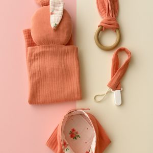 Organik Müslin Bebek Uyku Seti, pembe çiçek desenli pembe müslin örtü, uyumlu önlük, pembe müslin kundak ve mavi tavşan kulaklı ahşap diş kaşıyıcıdan oluşuyor.
